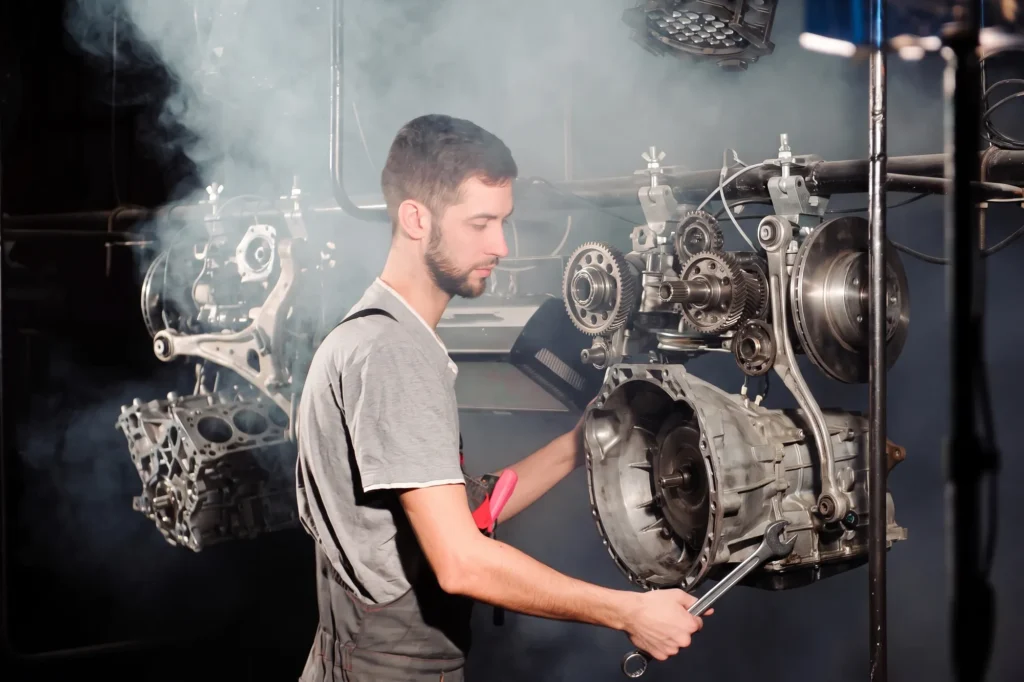 Technician with a spanner working on a gearbox and suspension parts mounted on an overhead rig, with fumes drifting through the workshop, representing automotive manufacturing applications for dust and fume extraction