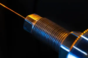laser beam striking the end of a threaded steel component against a dark background, representing laser shock peening of automotive parts