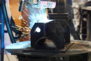 Robotic welding head joining a steel bracket on a turntable with a bright arc and a shower of sparks, representing welding of automotive parts