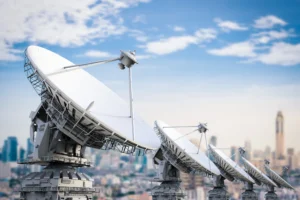 A row of large white satellite dish antennas on steel mounts pointing skyward, with a blurred city skyline and blue sky with clouds behind